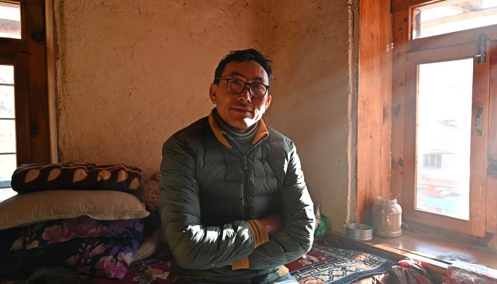 Man (Karma Sonam Tamang) sitting on bed by the window with his arms crossed, sun shining through the window on him.